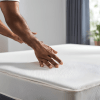 . A man is inspecting a mattress on a bed, his hand resting on the fabric to assess its softness and support.