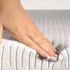 A hand gently touches the surface of a mattress, indicating softness and comfort.